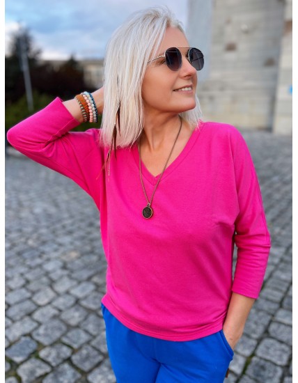 Kobieta w fuksjowej bluzce z długimi rękawami i niebieskich spodniach, stojąca na brukowanym placu.
