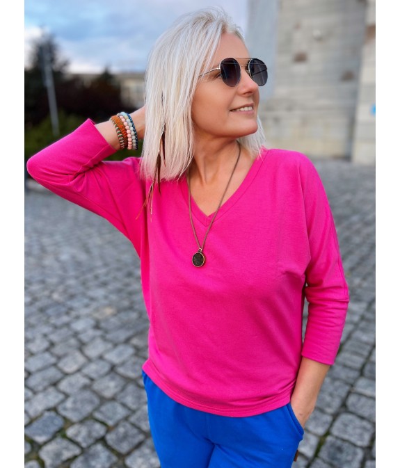 Kobieta w fuksjowej bluzce z długimi rękawami i niebieskich spodniach, stojąca na brukowanym placu.